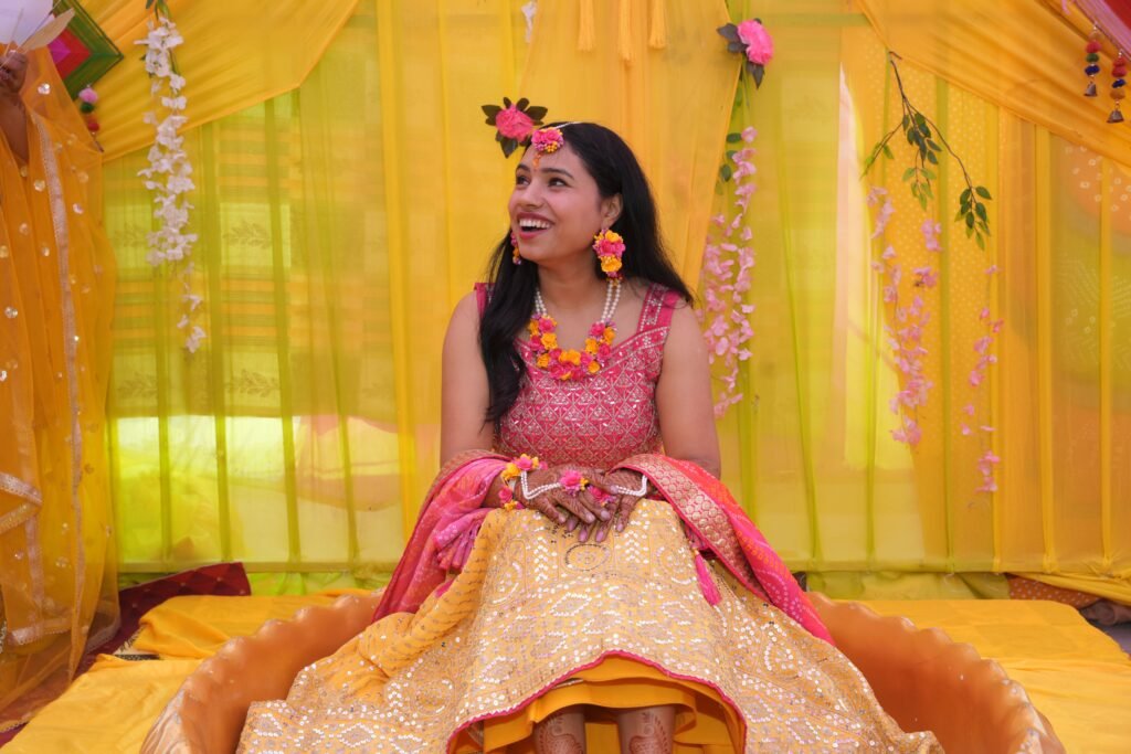 A happy bride at a colorful Indian haldi ceremony, adorned with flower jewelry, sitting joyfully.